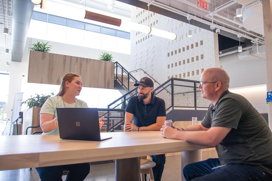 In a modern office setting, two men and woman with a laptop meet at a tall table.